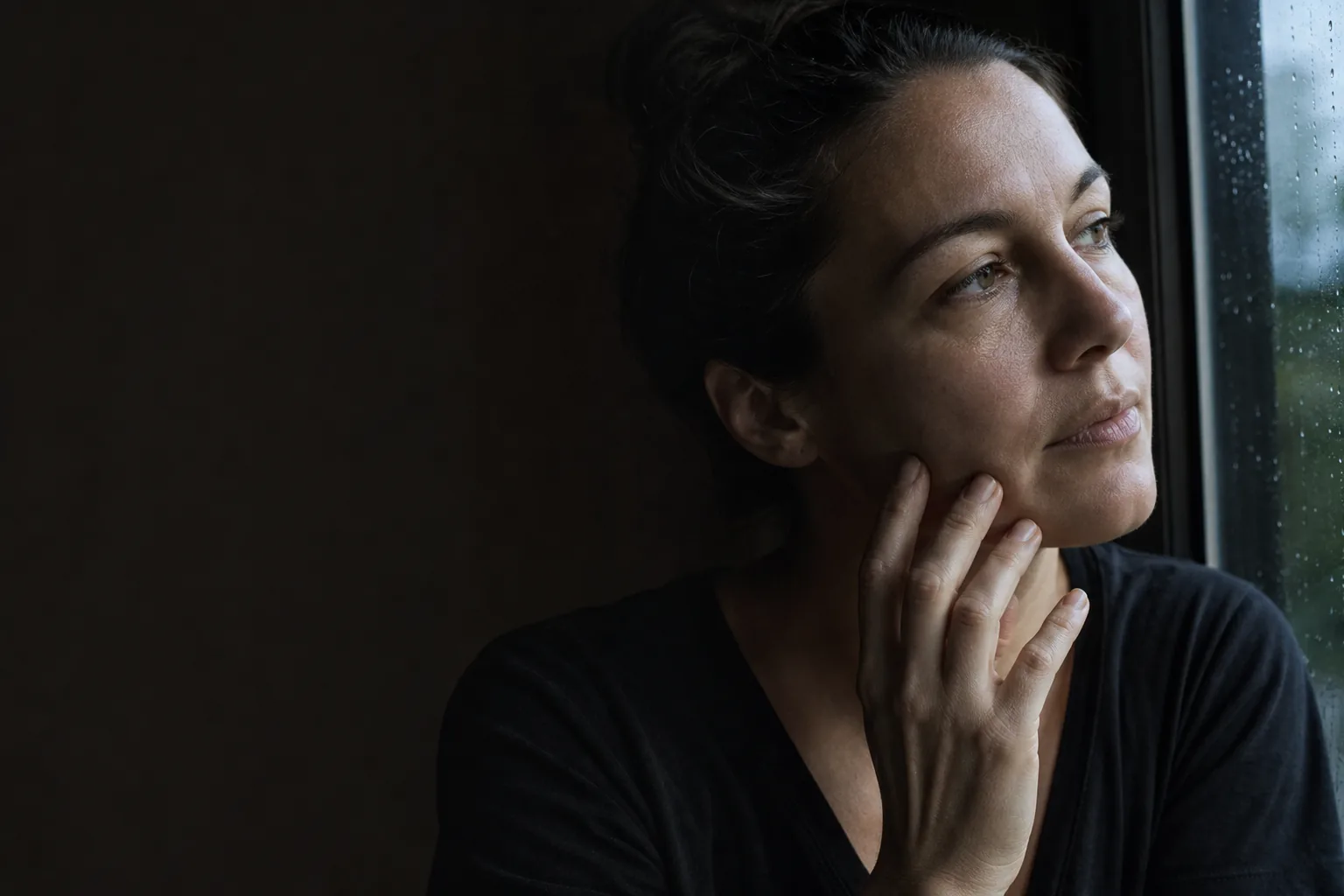 An adult pauses by a rain-darkened window with one hand resting at the jaw.